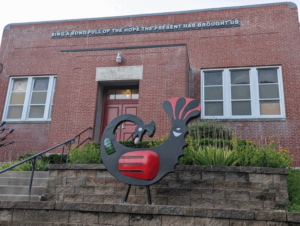 A brick building with a sign that says "Sing a Song Full of the Hope the Present has Brought Us." In front of the building stands a a sculpture of a Sankofa bird in red, black, and green.