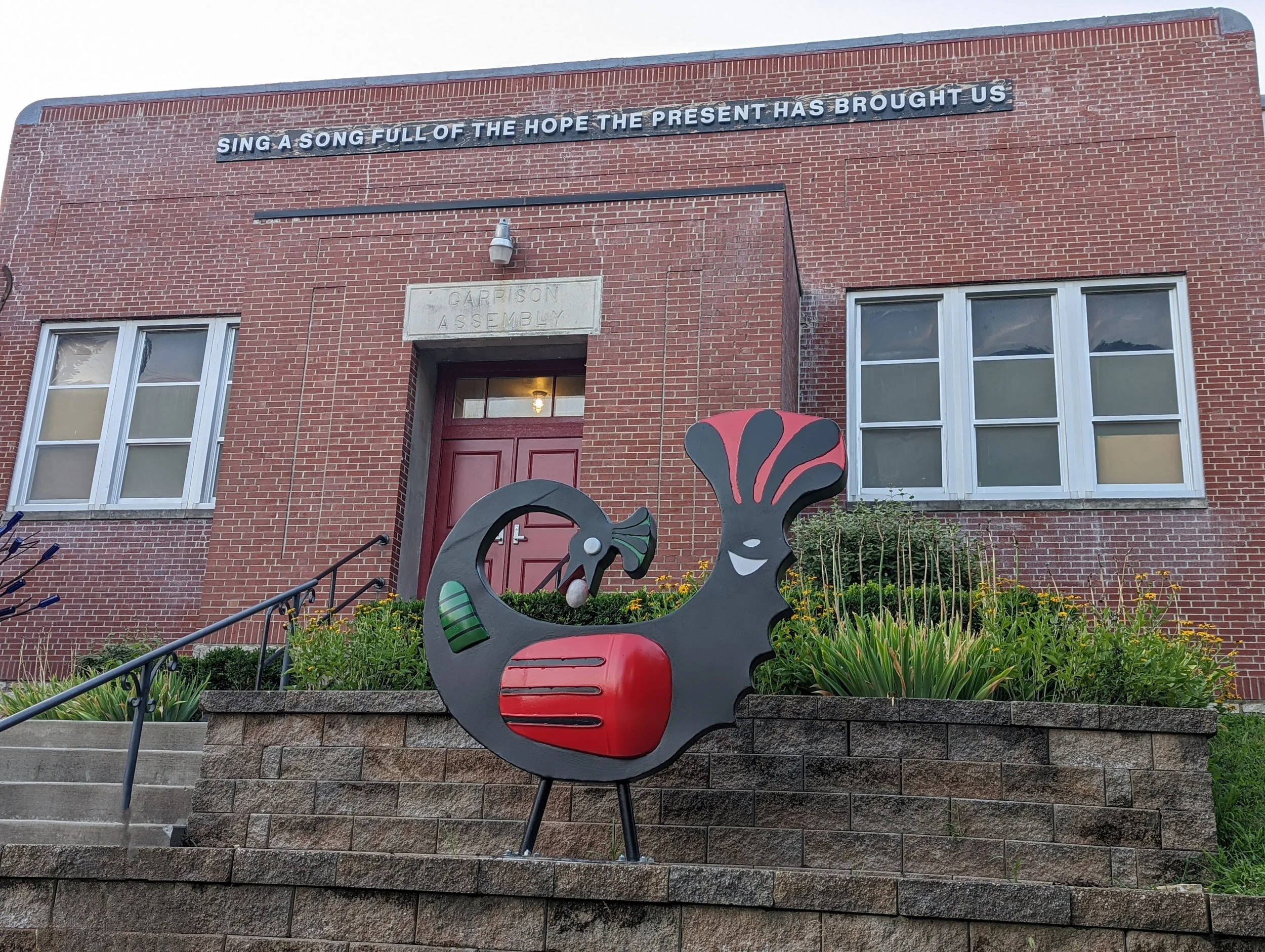 A brick building with a sign that says "Sing a Song Full of the Hope the Present has Brought Us." In front of the building stands a a sculpture of a Sankofa bird in red, black, and green.