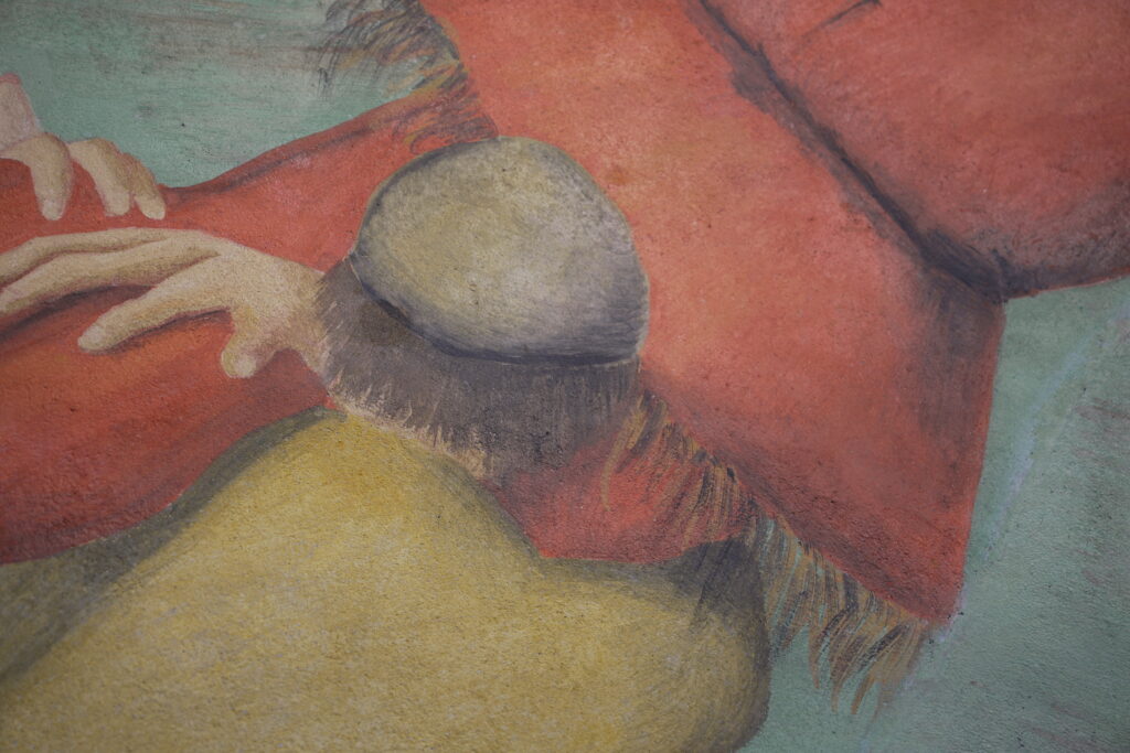 Section of mural with a man in a yellow shirt and a grey cap facing away from the audience. He holds a red scarecrow.