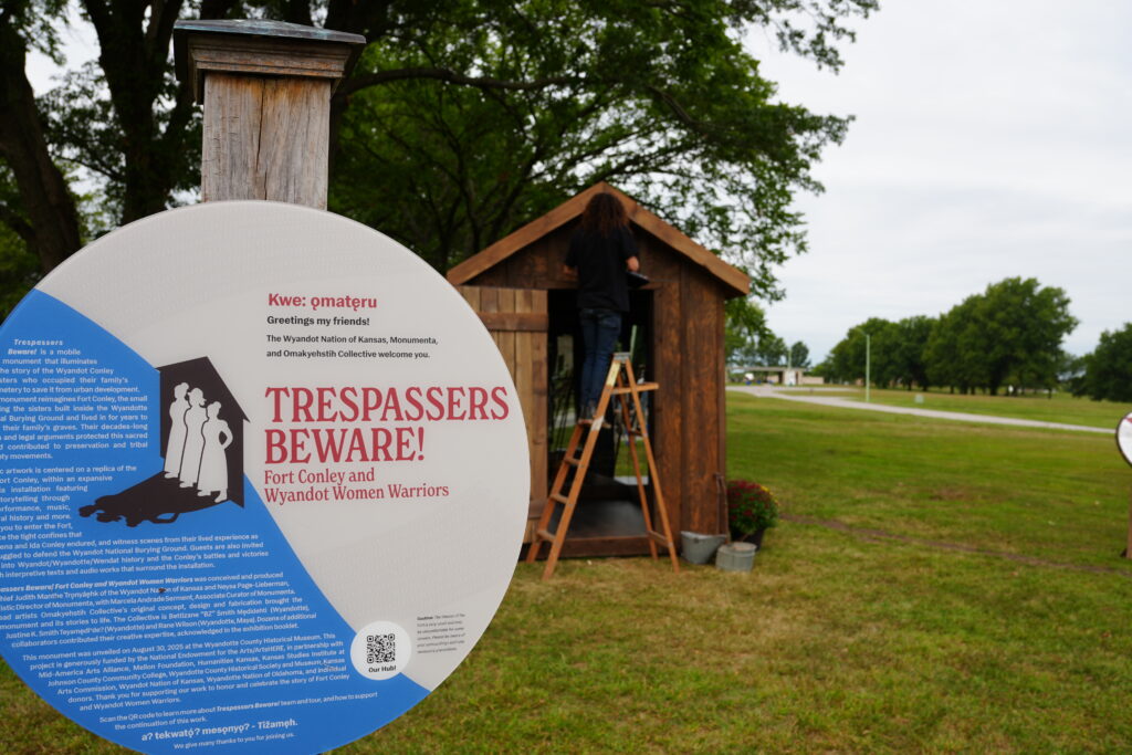 In the foreground, a sign says "Trespassers Beware! Fort Conley and Wyandot Women Warriors." In the background a small wooden hut sits in a grass field. A woman on a ladder works on its roof.