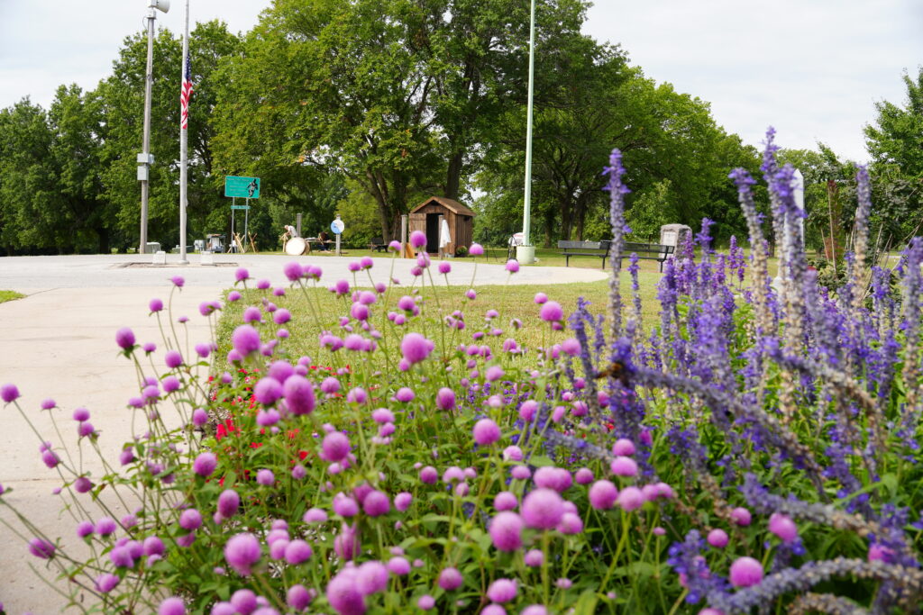 In the foreground, pink and purples flowers. In the background, flagpoles with the American flag, a concrete sidewalk, and a wooden hut.