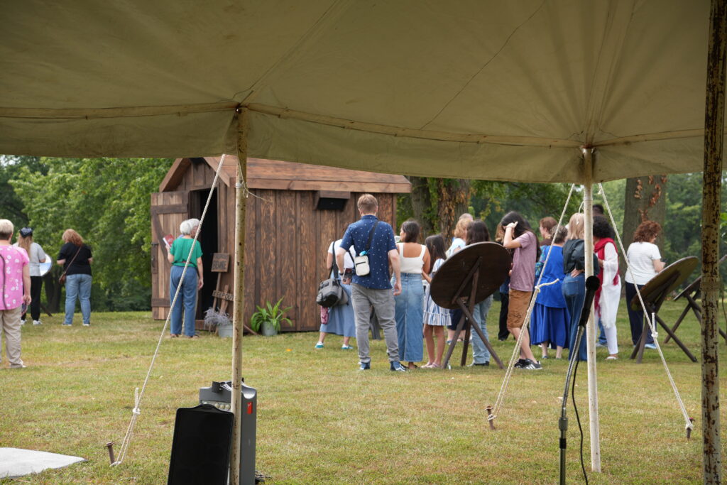 A crowd of people stand in front of a wooden hut. A canvas tent is in the foreground.