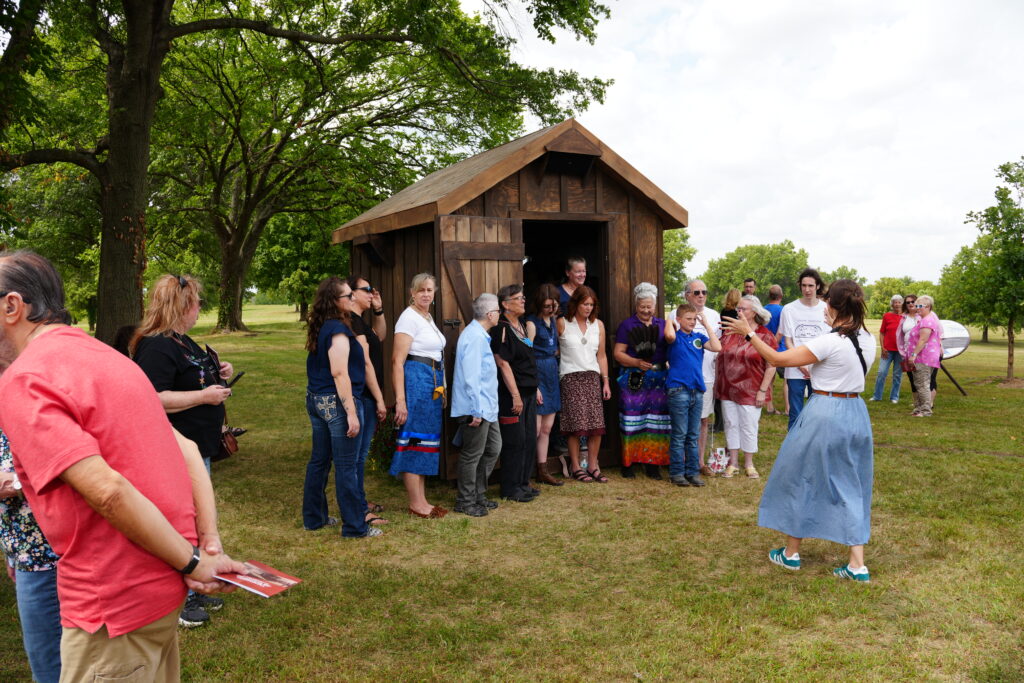 A group od people pose in front of a wooden hut. A photographer directs them to stand closer together.