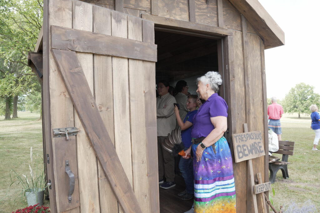A women with grey hair, a purple shirt, and a colorful skirt stands in the doorway of a wooden hut. A sign outside the hut reads "Trespassers Beware." Another reads "Keep out"
