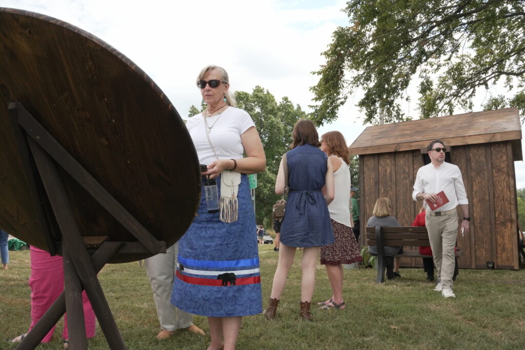 A woman stands and reads from a sign. In the background five people stand around a wooden hut.