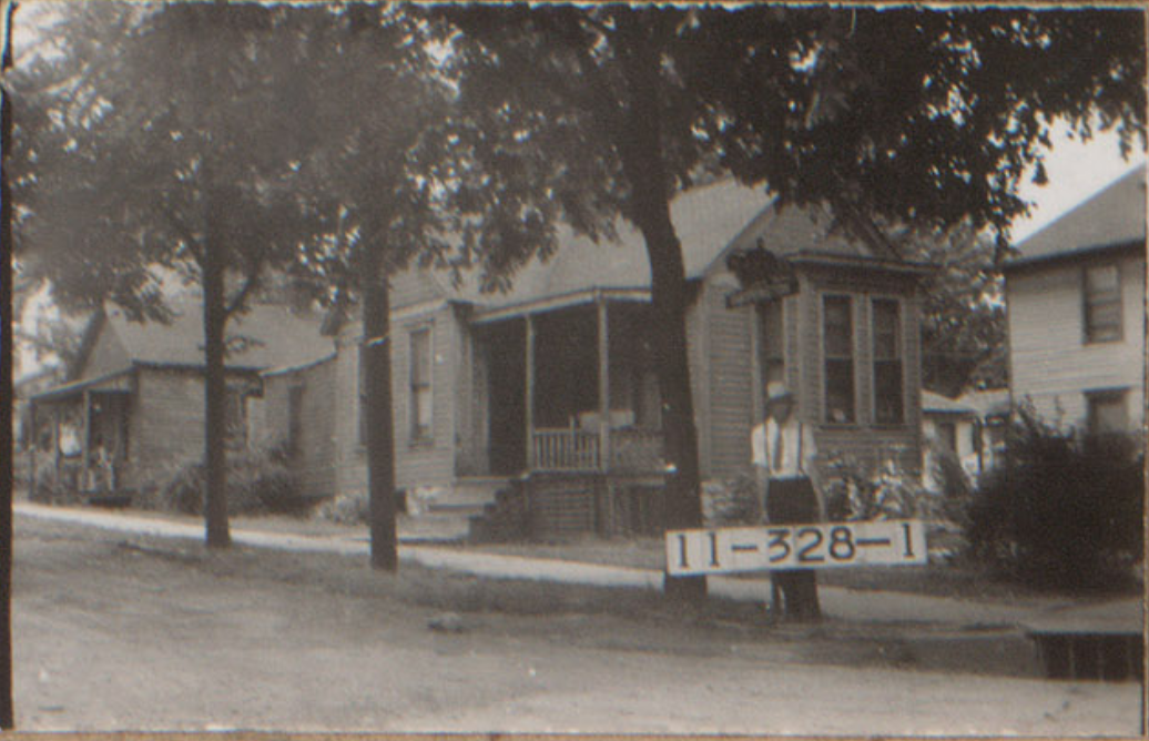 1940 tax assessment photo of a man standing holding a tax sign reading 11-328-1. He stands on a tree lined street in front of 3 houses.