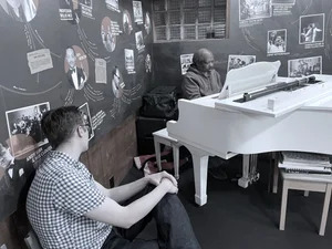 A man sits next to another man playing a white piano. Behind them, a wall with photos of jazz musicians fills the room.