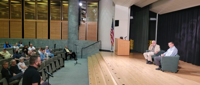 Man stands at microphone in front of auditorium, asking 2 men on stage a question